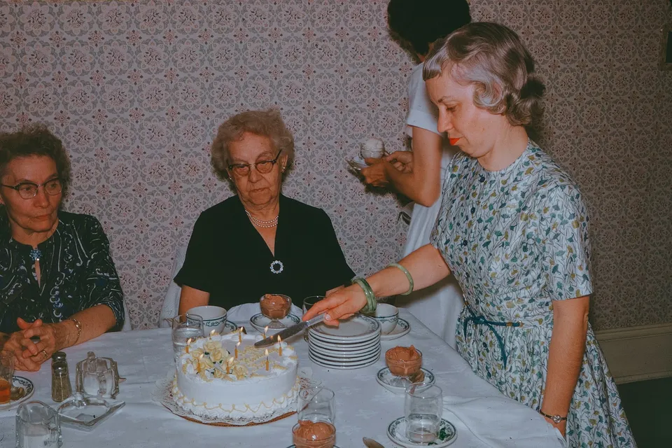 Una mujer corta la tarta de cumpleaños de una señora mayor sentada a la mesa, en una foto antigua de una celebración familiar