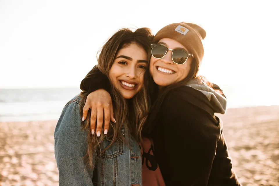 Dos amigas abrazadas y sonriendo en una playa, con el sol de frente