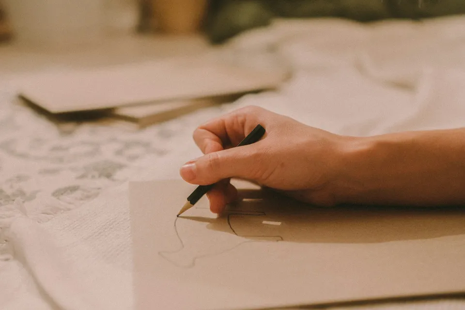 Una mano escribe con bolígrafo en una tarjeta de papel kraft, sobre un mantel de encaje