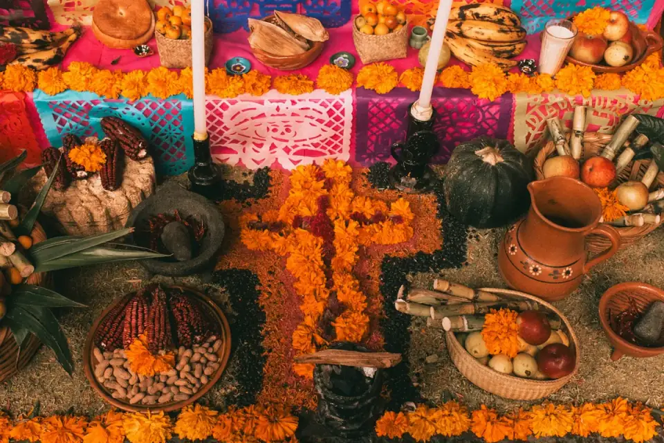 An ofrenda on the floor: a path of marigold petals, candles, a clay jug, corn, fruit and a squash over papel picado