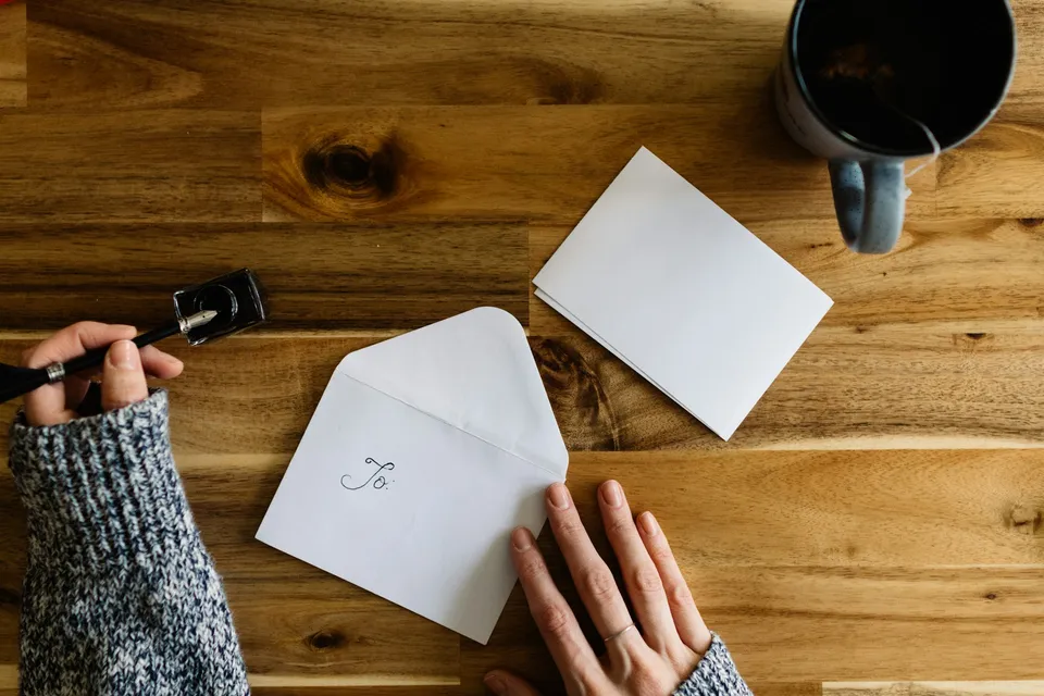 Hands writing on an envelope at a wooden table, next to a cup of coffee