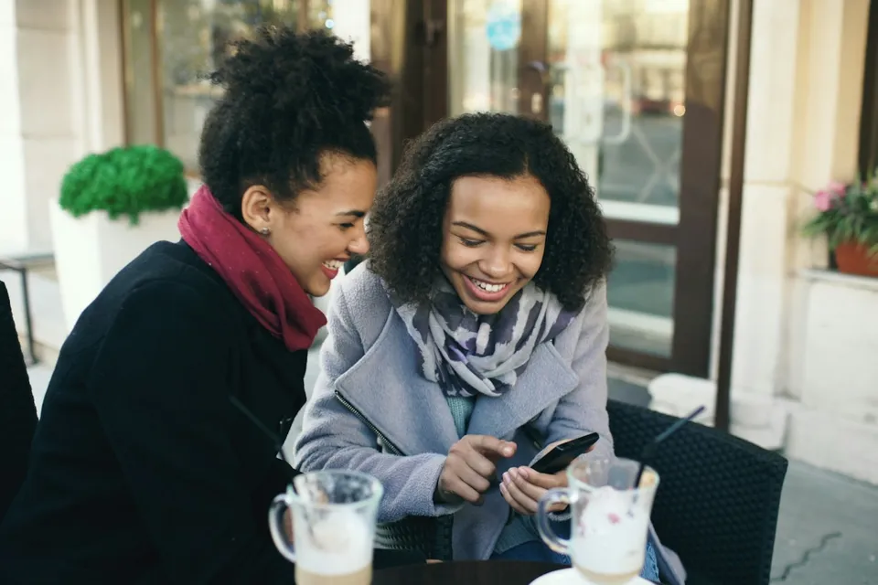 Two friends laughing together while looking at a phone at a café
